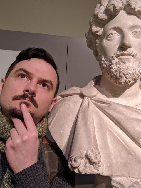 Brandon McPeak in 3/4 profile beside a marble bust of Marcus Aurelius at the British Museum.